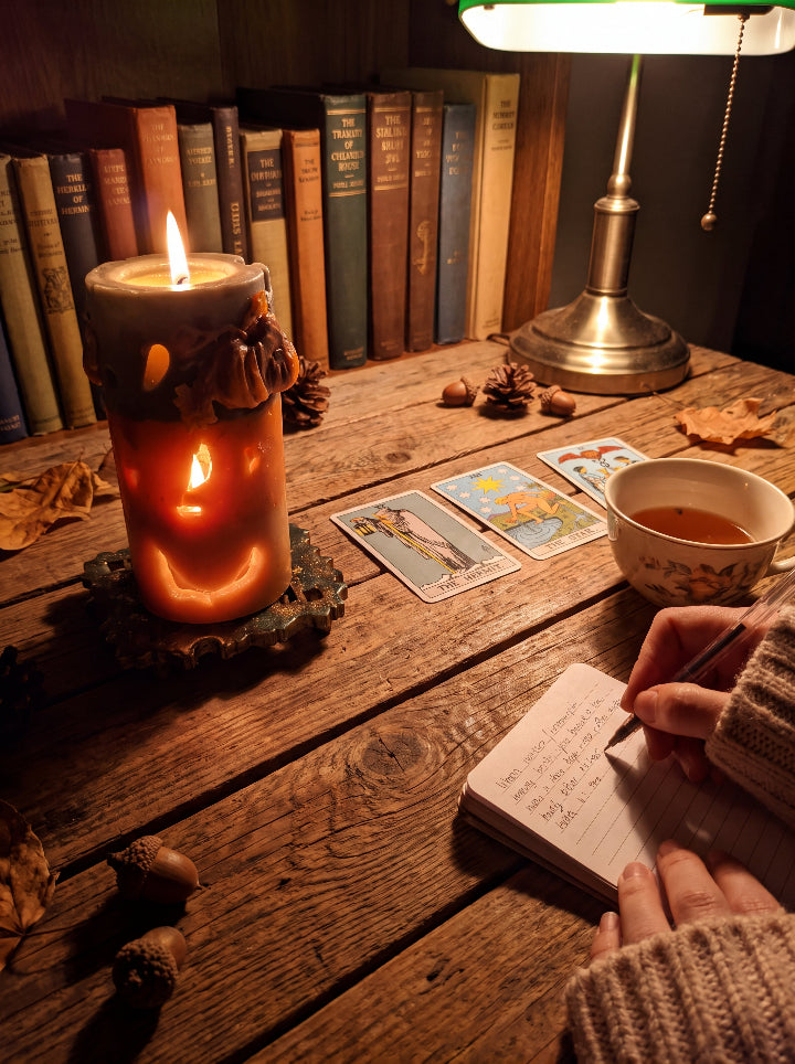 Lit Witch’s Harvest candle beside tarot cards, tea and a handwritten journal on a wooden desk.
